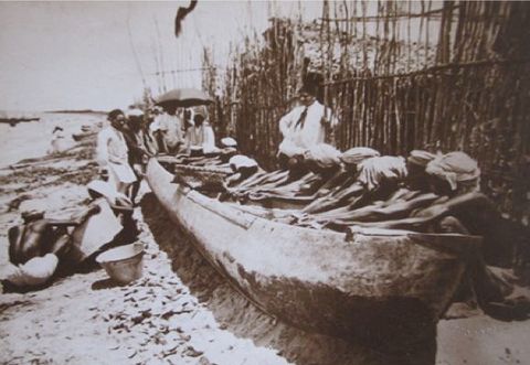 Black and white photograph. On a sandy beach, dark-skinned men with light cloths wrapped around their heads sit alongside a long, wooden boat. Their arms stretch in front of them, engaged in a task hidden by the boat. Men in white clothing look on, some under a parasol at the far end of the boat.