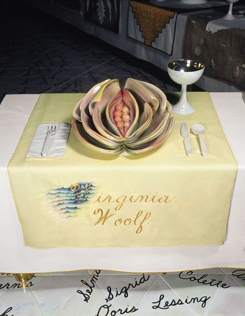 Installation. A place setting at a table has a highly sculptural plate with deep folds around a narrow opening in shades of pale pink, yellow and green. The yellow cloth beneath this setting is embroidered with gold letters, \“Virginia Woolf\”.